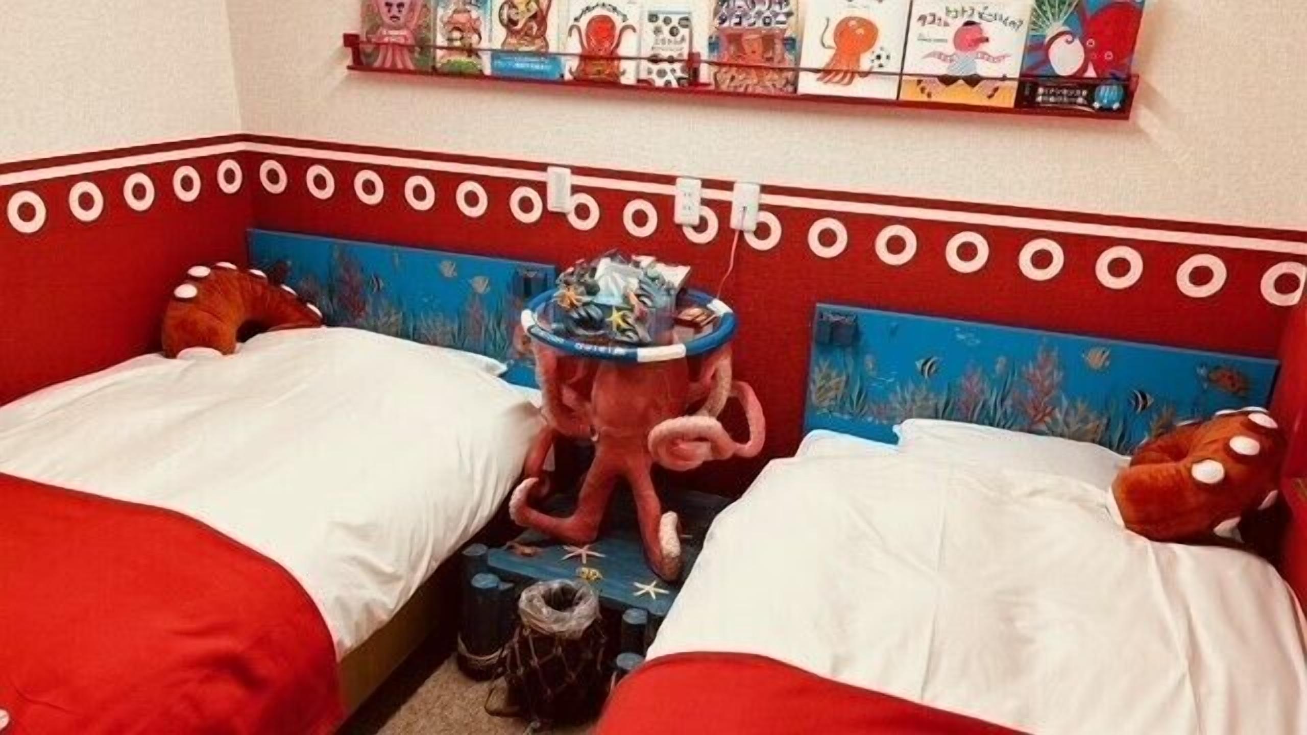 Un dormitorio con dos camas, cada una con ropa de cama roja y cabecero azul. Encima de las camas hay una estantería con varios libros y un juguete.
