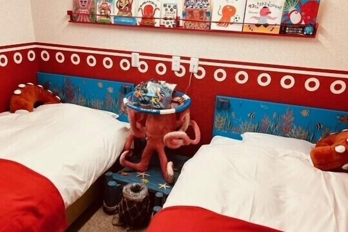 Un dormitorio con dos camas, cada una con ropa de cama roja y cabecero azul. Encima de las camas hay una estantería con varios libros y un juguete.
