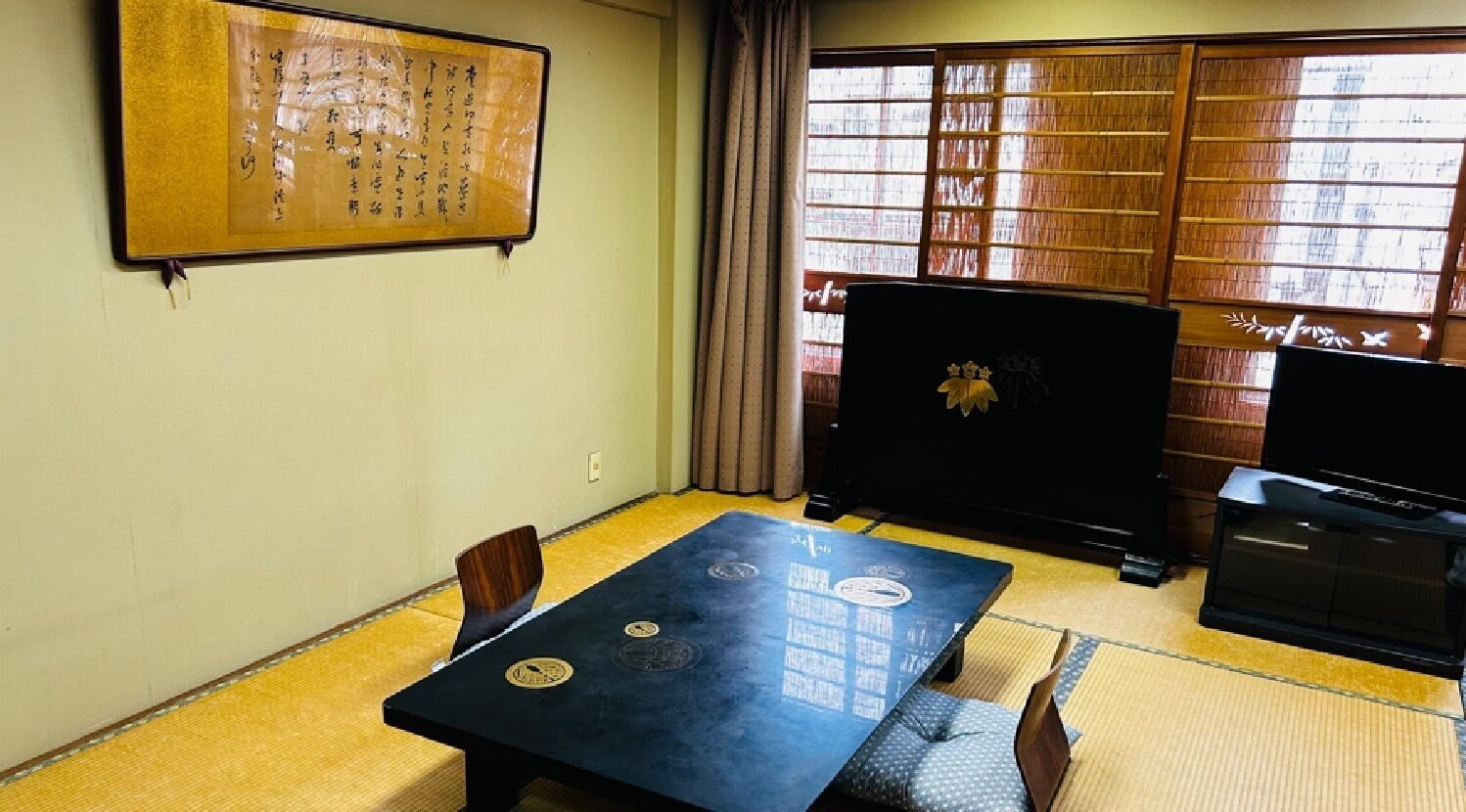 Una habitación con suelo de tatami, una mesa baja con cojines, un televisor y una obra de caligrafía japonesa tradicional en la pared.