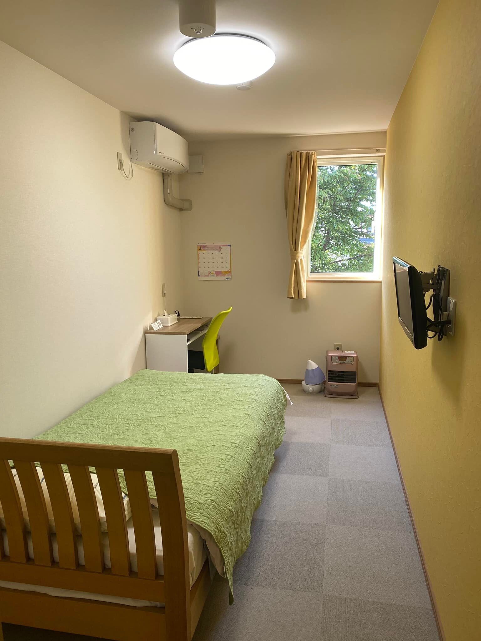 Quarto pequeno e bem iluminado, com cama, escrivaninha, cadeira e janela com cortinas.