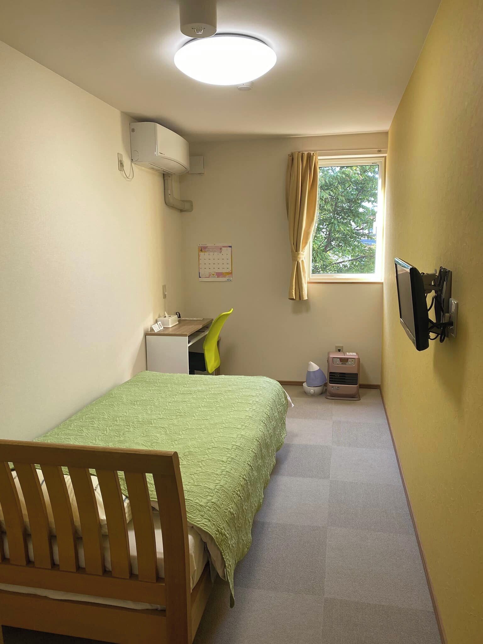 Ein kleiner, gut beleuchteter Raum mit Bett, Schreibtisch, Stuhl und einem fenster mit Vorhängen.