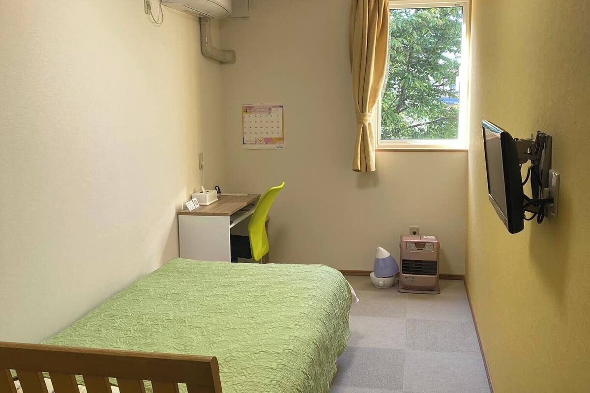 Una habitación pequeña y bien iluminada con cama, escritorio, silla y ventana con cortinas.