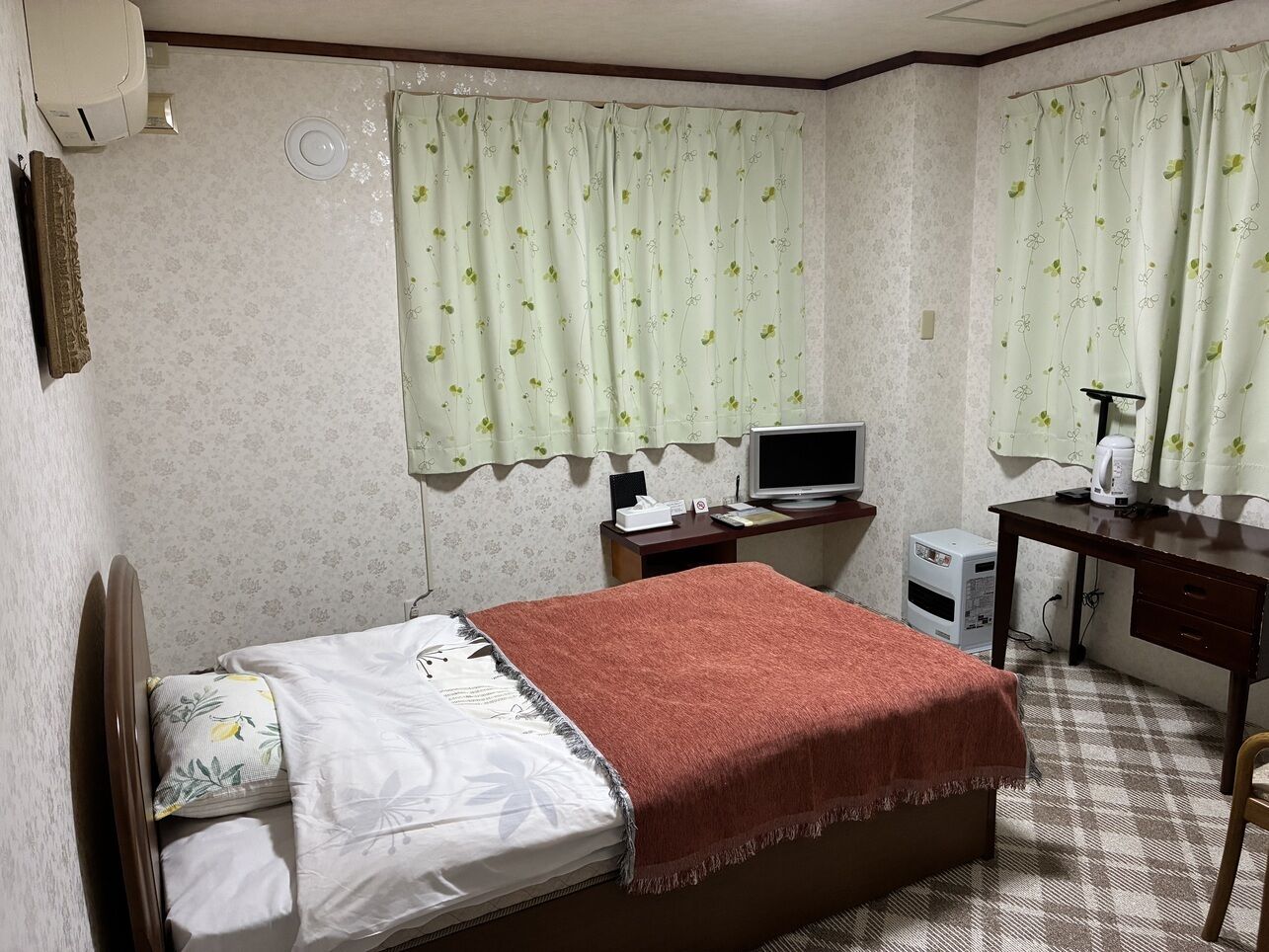 Quarto com cama, escrivaninha, cadeira, televisão e aquecedor pequeno.