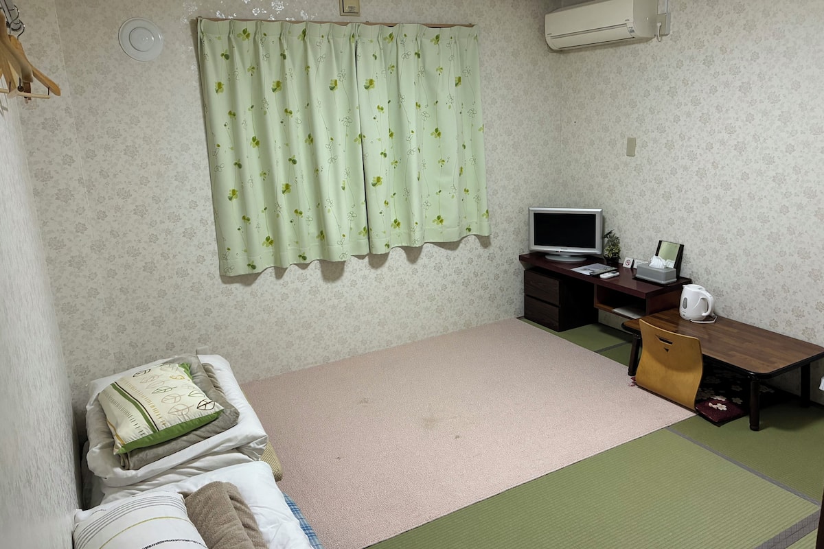 Una habitación pequeña y ordenada con una cama, un escritorio con computadora, televisión y aire acondicionado.