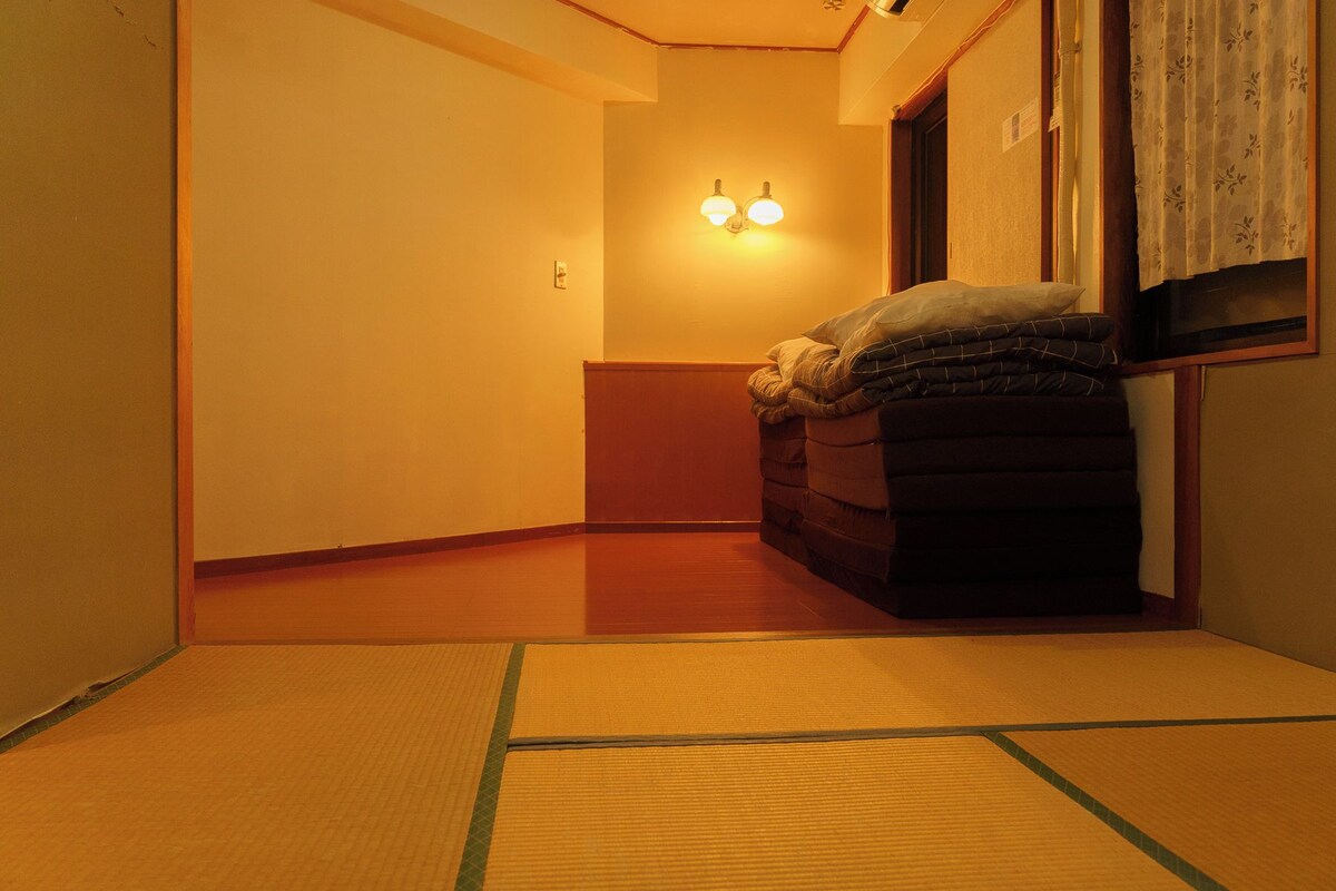 A narrow hallway with tatami mats, a bed with a blanket, and a wall-mounted light fixture.