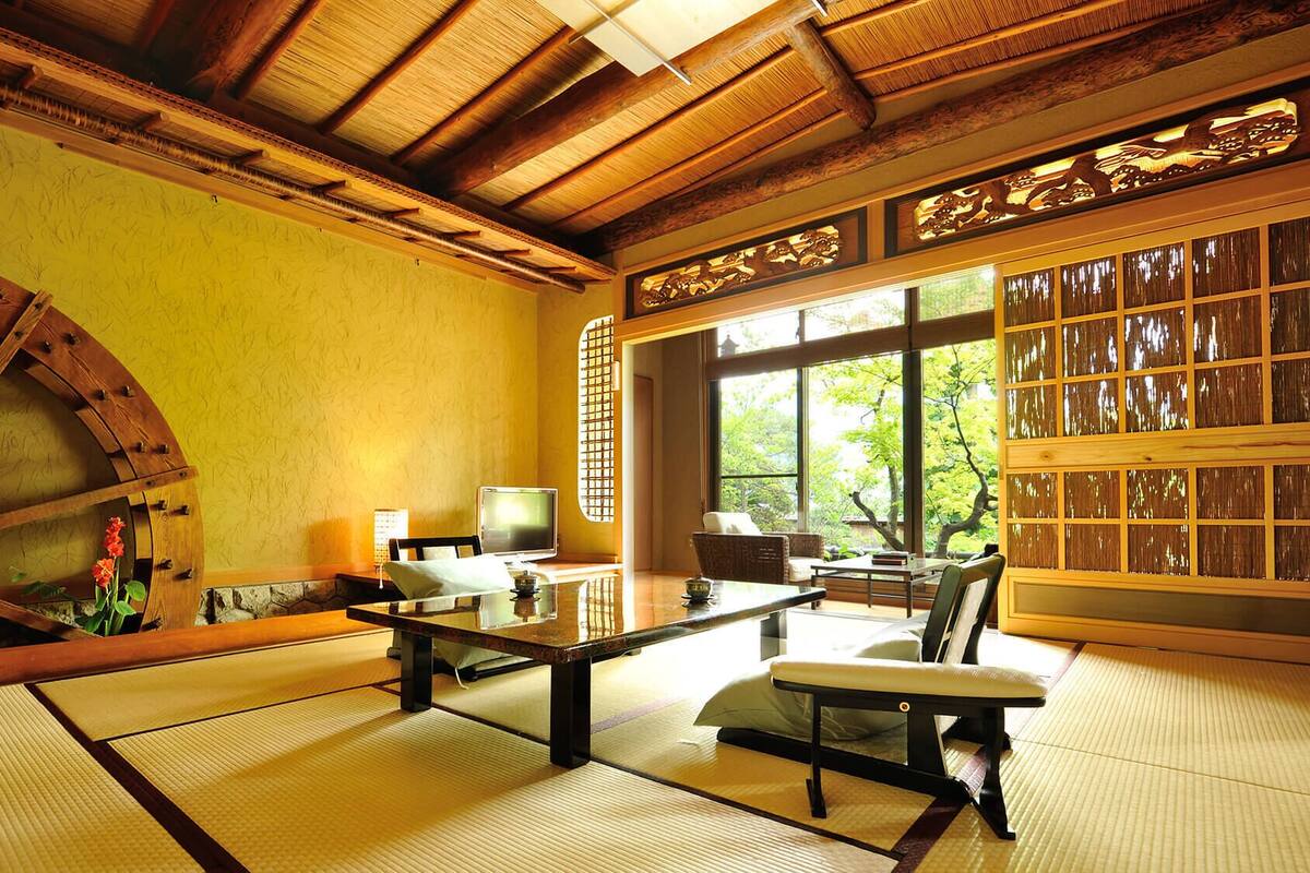 A traditional Japanese room with tatami flooring, a wooden ceiling, a large wooden wheel, and a view of greenery through large windows.