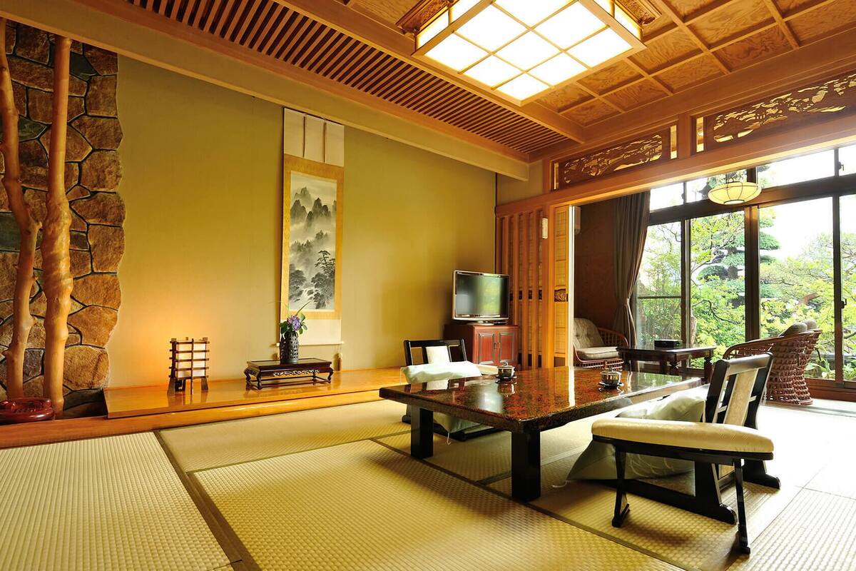 A traditional Japanese-style room with tatami flooring, a low table, a television, and a view of greenery outside.