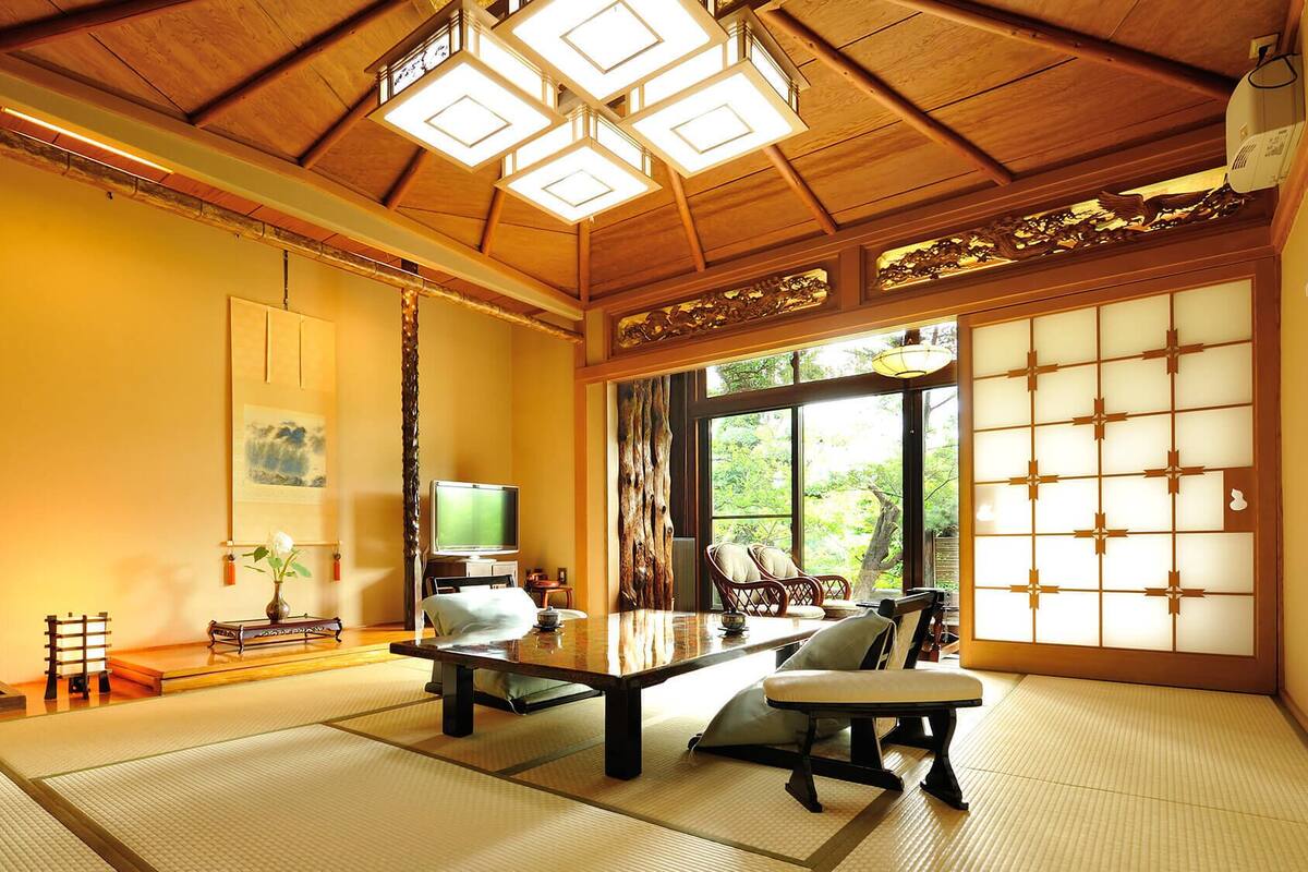 A traditional Japanese room with tatami flooring, a low table, a television, and a view of greenery outside.