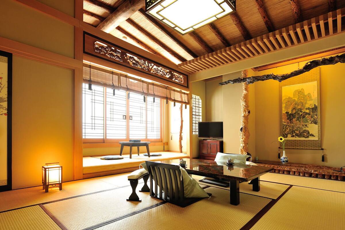 A traditional Japanese room with tatami flooring, a tatami mat table, a low wooden bench, a television, and a painting.