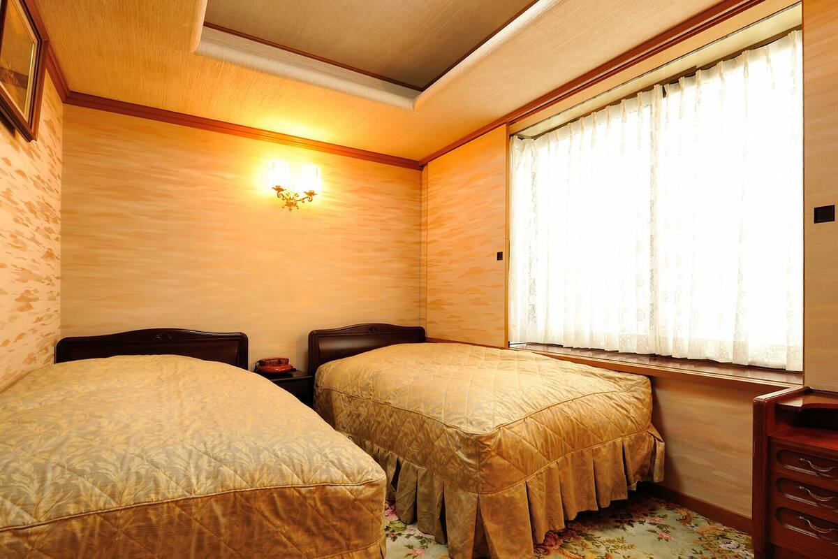 A small bedroom with two beds, a nightstand, a lamp, a window with curtains, and a dresser.