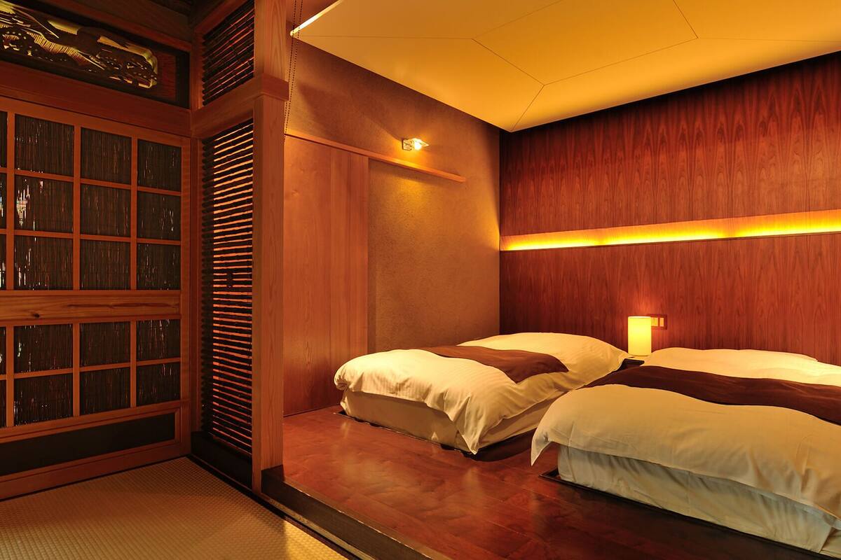 A hotel room with two beds, wooden paneling, a large window with blinds, and a wall-mounted light fixture.