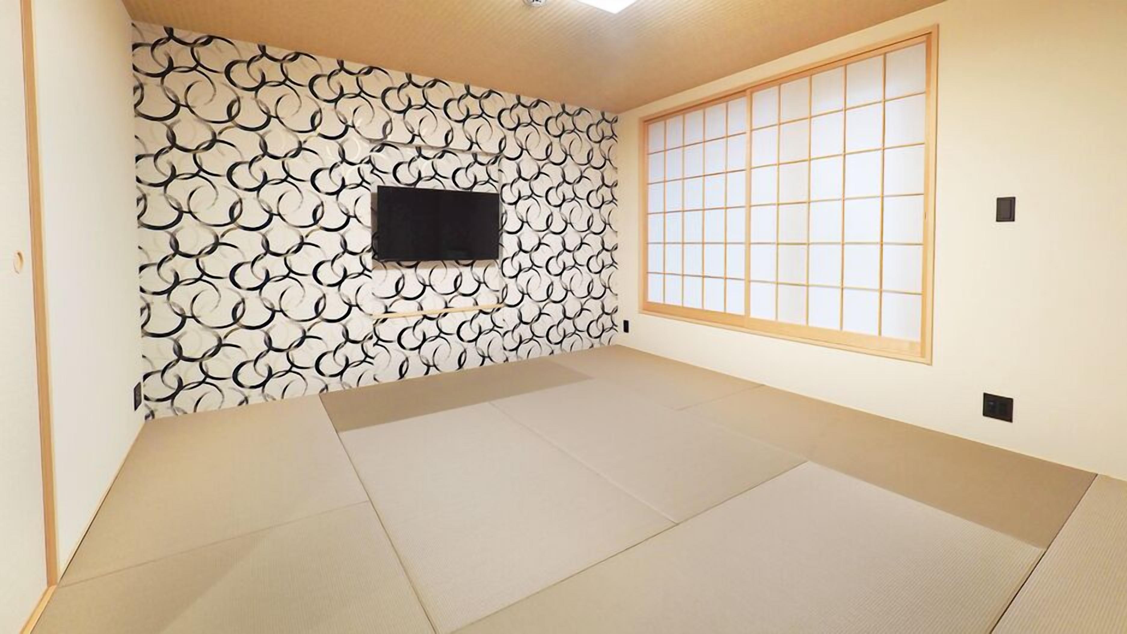 Quarto com piso de tatame, uma TV preta fixada numa parede estampada e uma porta de papel shoji.