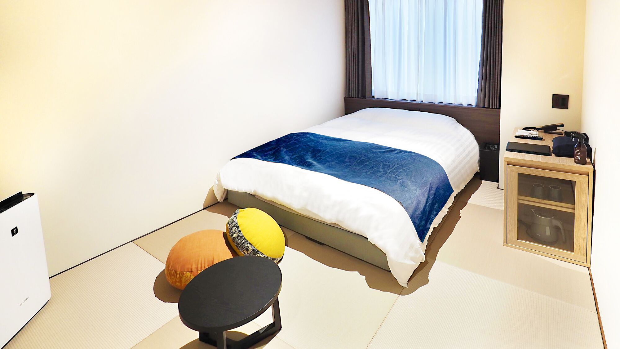 Quarto de hotel com cama, mesinha, cadeira, minibar e janela com cortinas.