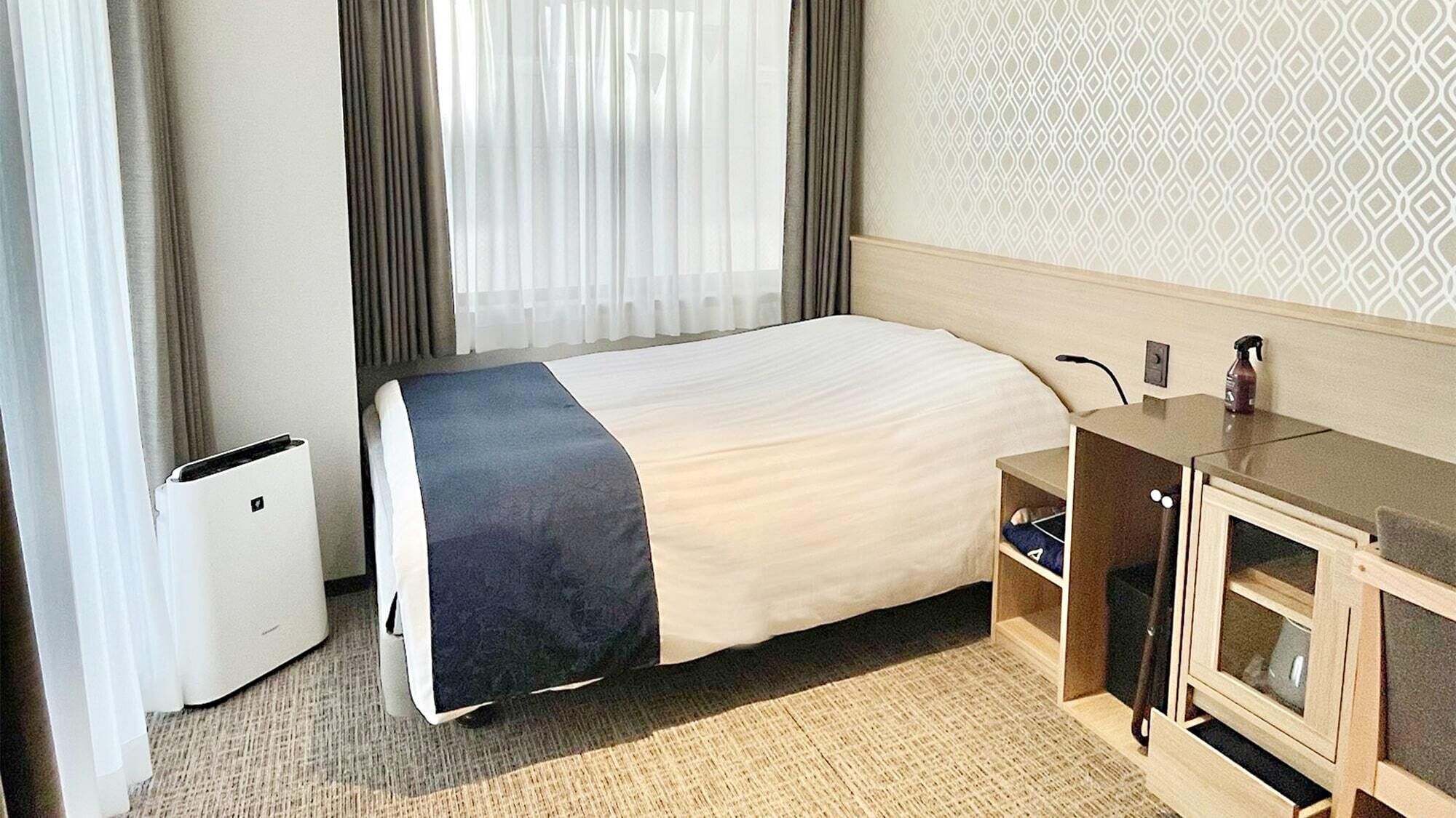 Quarto de hotel com uma cama, escrivaninha, cadeira, armário pequeno e lixeira.