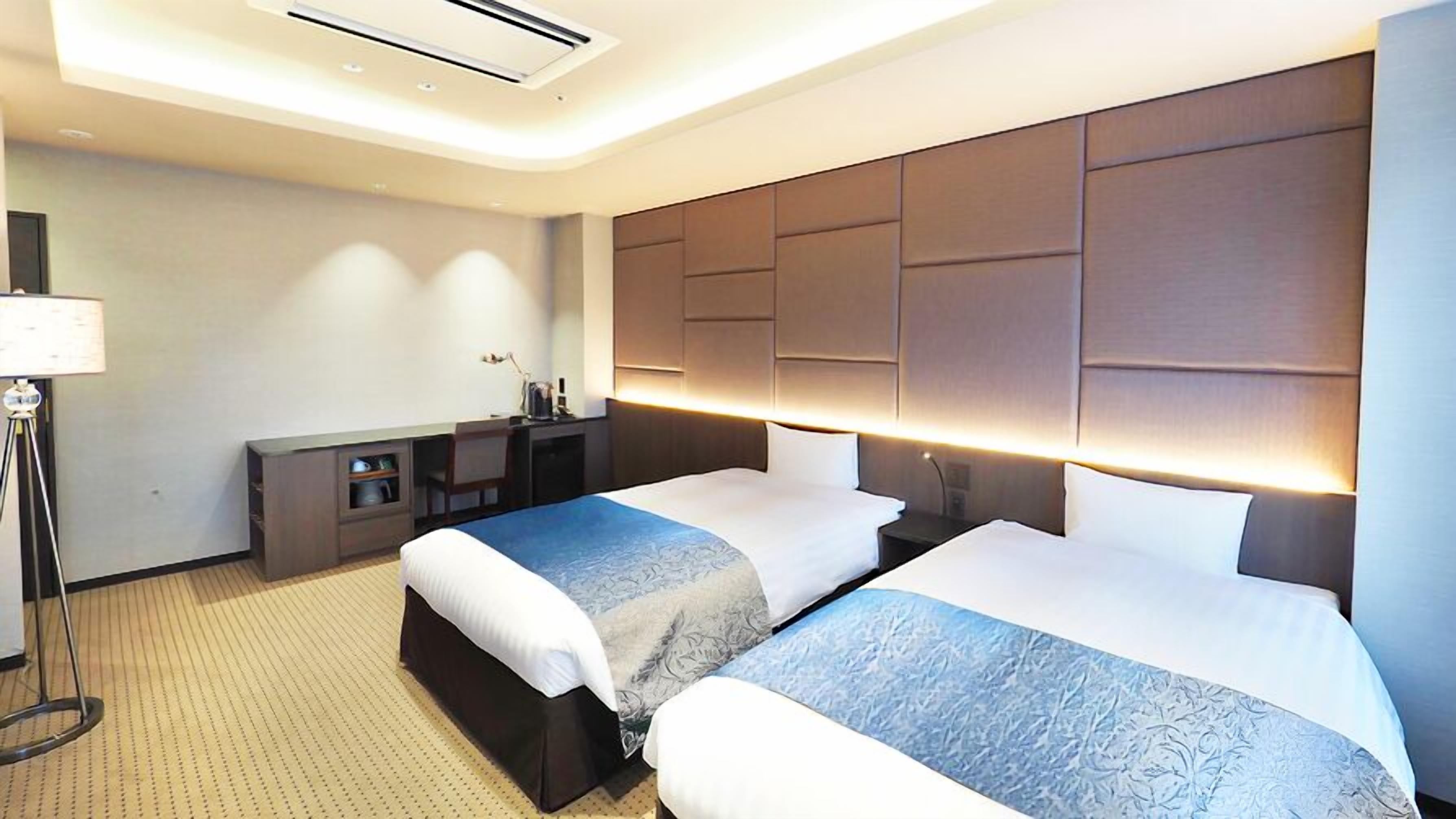 Quarto de hotel com duas camas, uma escrivaninha, uma luminária e um respiradouro no teto.