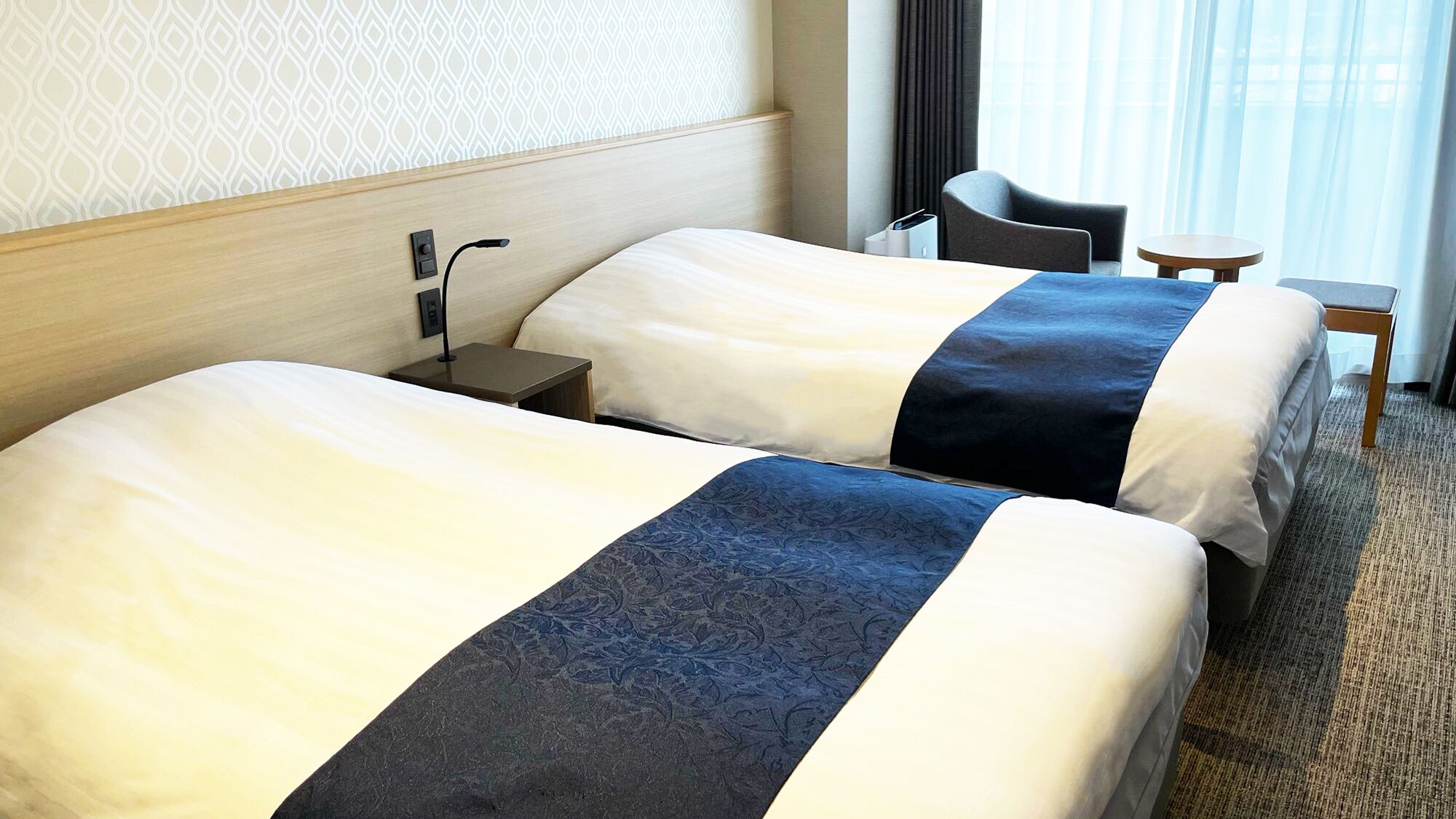 Una habitación de hotel con dos camas, una mesita de noche, una silla, una pequeña mesa y un ventanal con cortinas.
