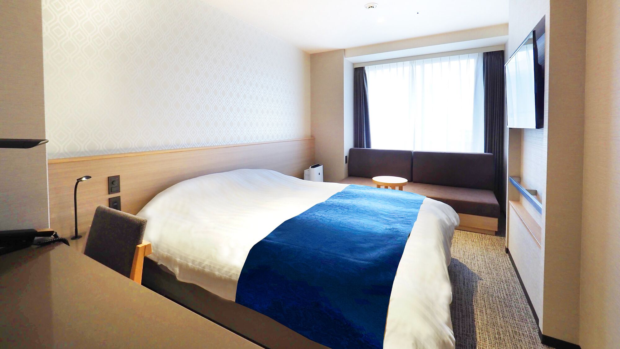 Una habitación de hotel con cama, una mesita, un sofá, una lámpara y una ventana con cortinas.