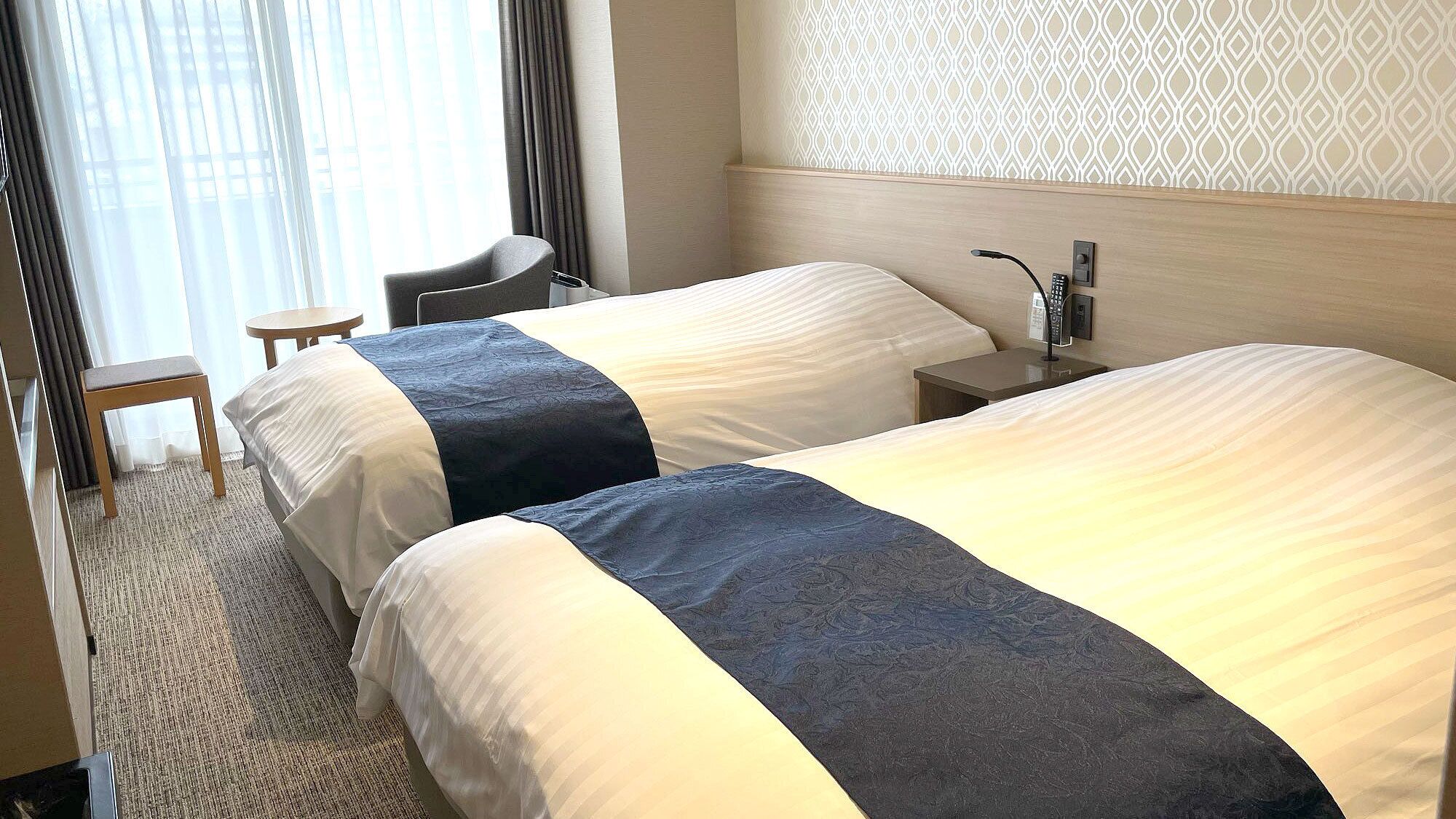 Quarto de hotel com duas camas, uma escrivaninha, uma cadeira e um papel de parede estampado.