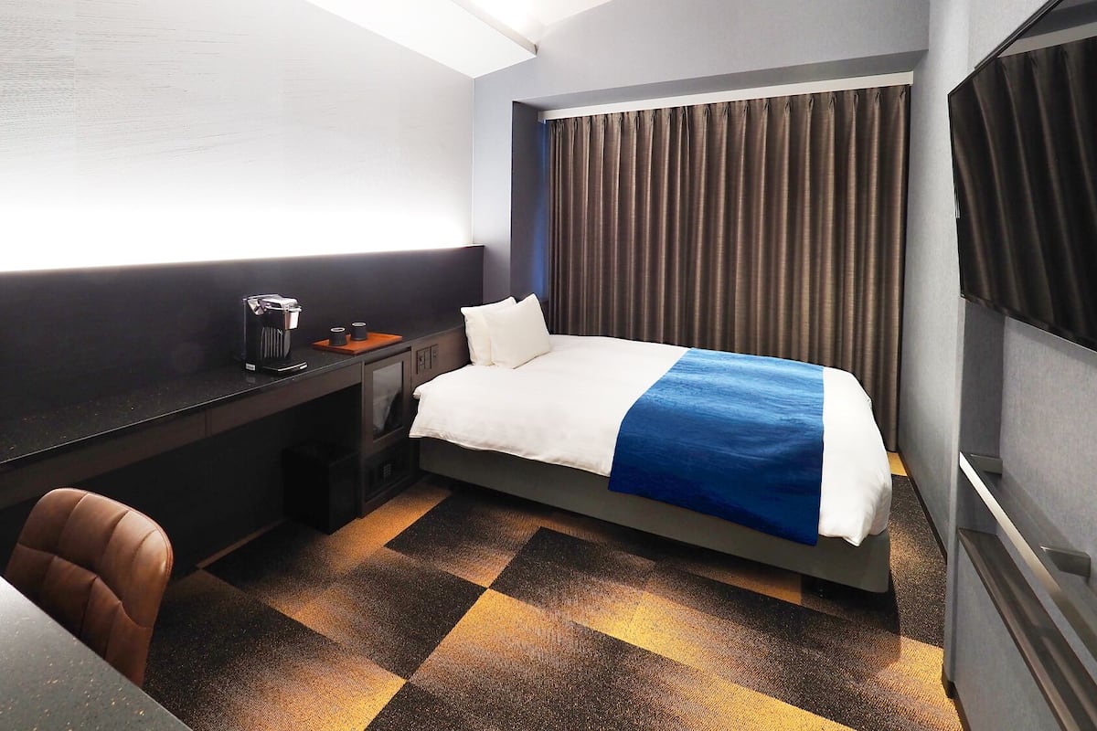 Habitación de hotel con cama, escritorio, silla, cafetera y ventana con cortinas.