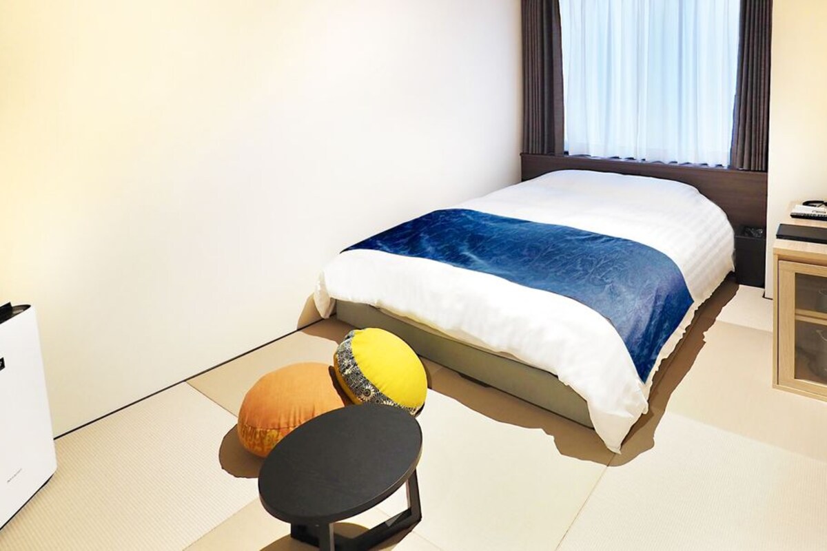 Hotellihuone, jossa on sänky, pieni pöytä, tuoli, yöpöytä lampun kanssa ja ikkuna, jossa on verhot.