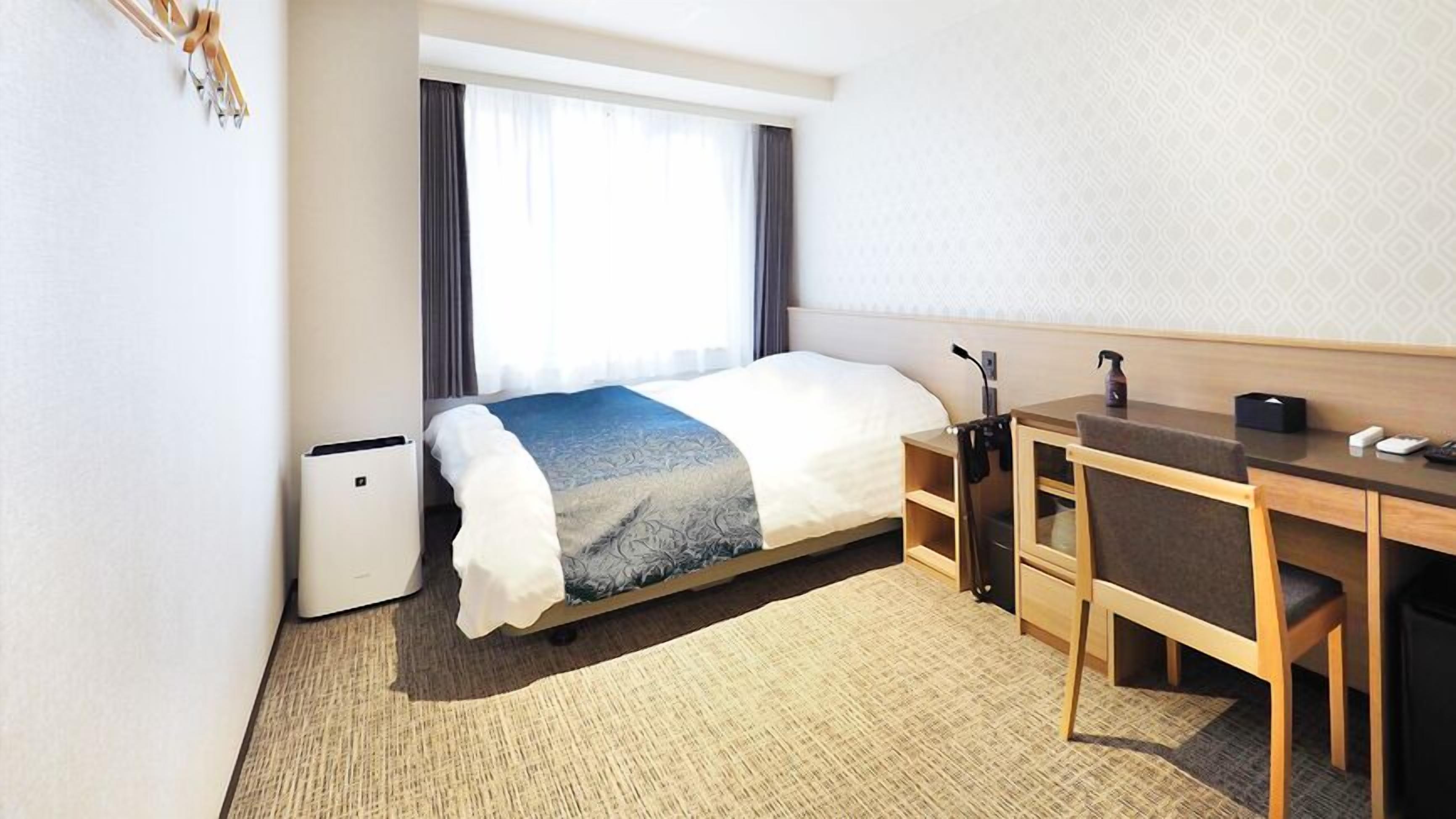 Quarto de hotel com cama, escrivaninha, cadeira e um pequeno aparelho de ar condicionado.