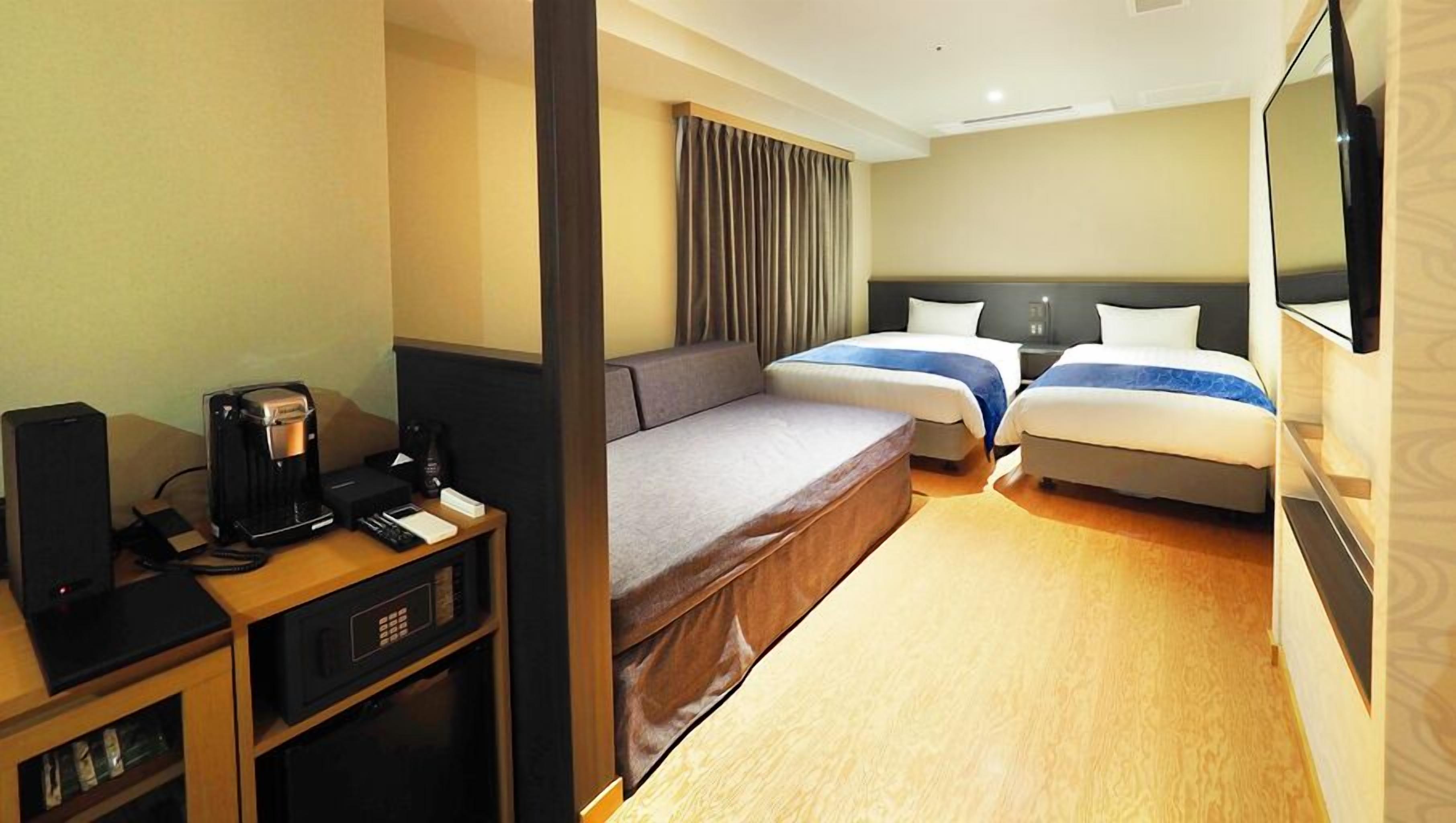Quarto de hotel com escrivaninha, duas camas, um sofá, televisão e cafeteira.
