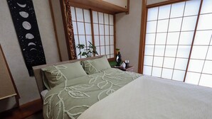 A traditional Japanese-style room with tatami flooring, a low bed with a green and white patterned bedspread, a shoji screen window, and a small bedside table with a plant and a decorative item. - Greenmist Atami Shizenkyo (Atami)