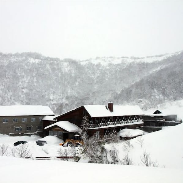 Un chalet enneigé, composé d’un grand bâtiment et d’une construction plus petite, entouré d’arbres et de collines.