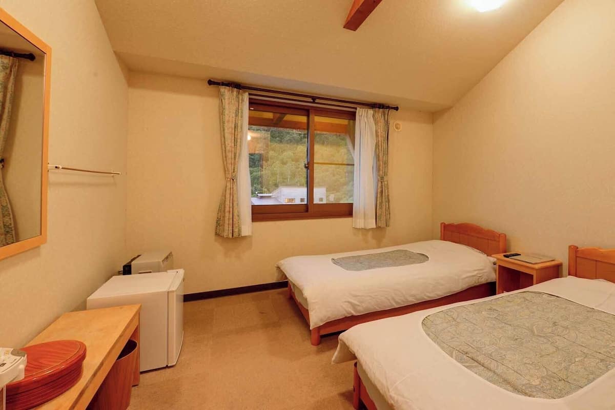 Un dormitorio con dos camas, una mesita de noche, un mueble auxiliar, una ventana con cortinas y un espejo en la pared.