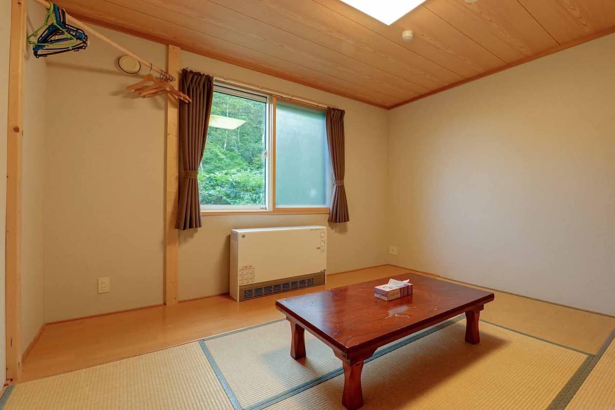 Una habitación con techo de madera, ventana con cortinas, radiador, suelo de esterilla tatami y una mesa de madera con una caja de pañuelos.