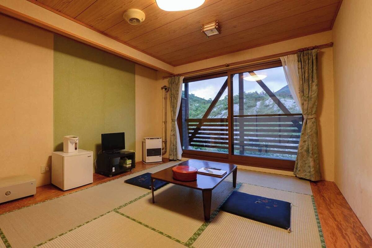 Una habitación con suelo de tatami, televisor, calentador y ventana con vista a una estructura de madera.