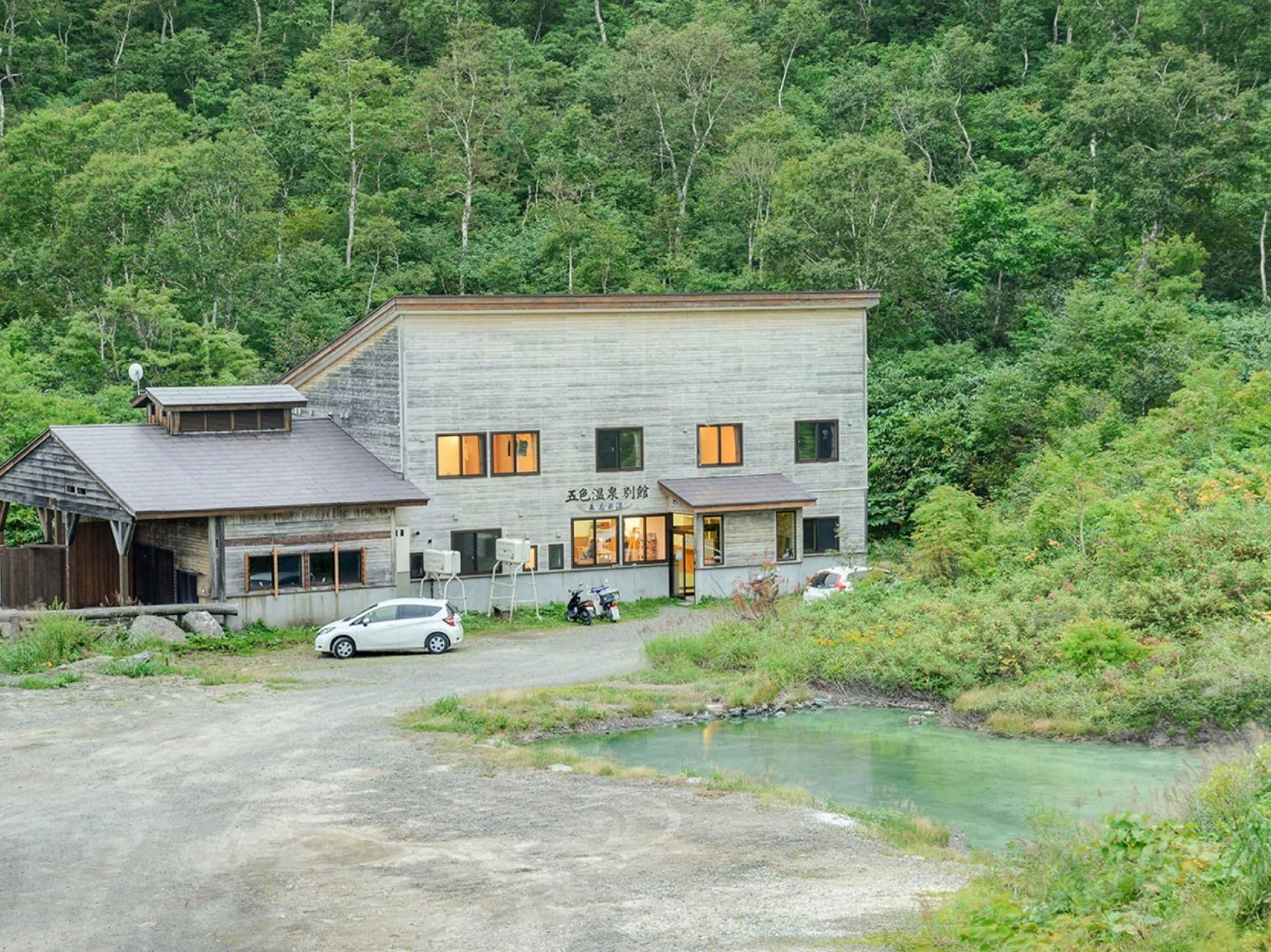 A two-story building with a flat roof, a car parked in front, and a small body of water nearby.