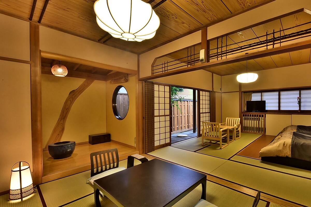 A traditional Japanese-style room with tatami flooring, a low table, chairs, a bed, and a television.