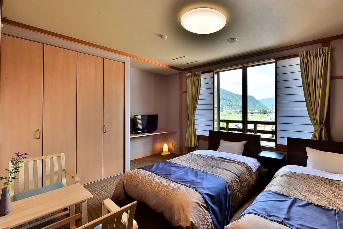 A hotel room with two beds, a TV, a desk, and a window with curtains.