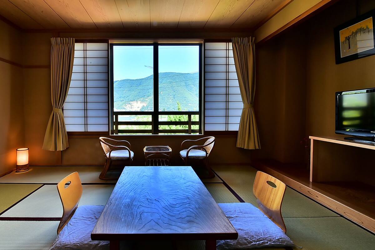 A traditional Japanese room with tatami flooring, a low table with cushions, a television, and a view of a mountainous landscape through shoji screens.