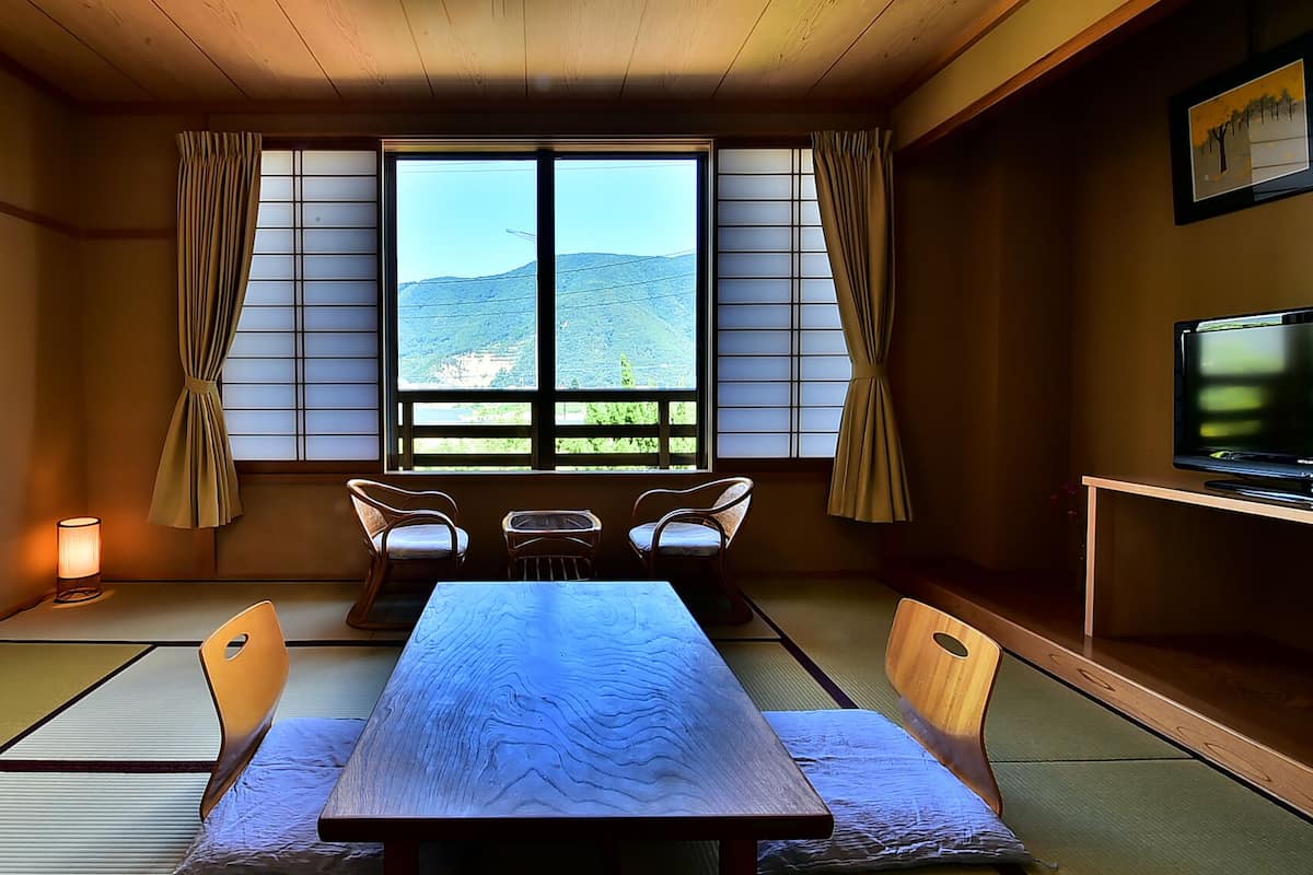 A traditional Japanese room with tatami flooring, a low table with cushions, a television, and a view of a mountainous landscape through shoji screens.