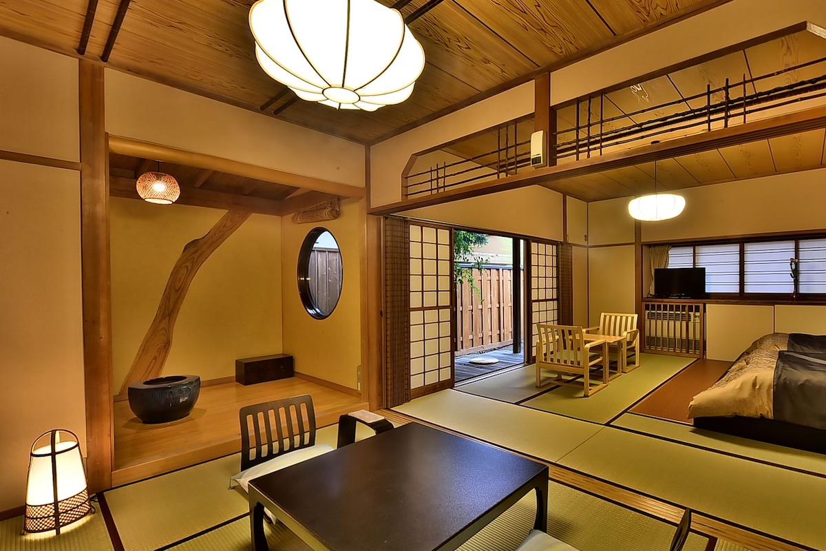 A traditional Japanese-style room with tatami flooring, a low table, chairs, a bed, and a television.