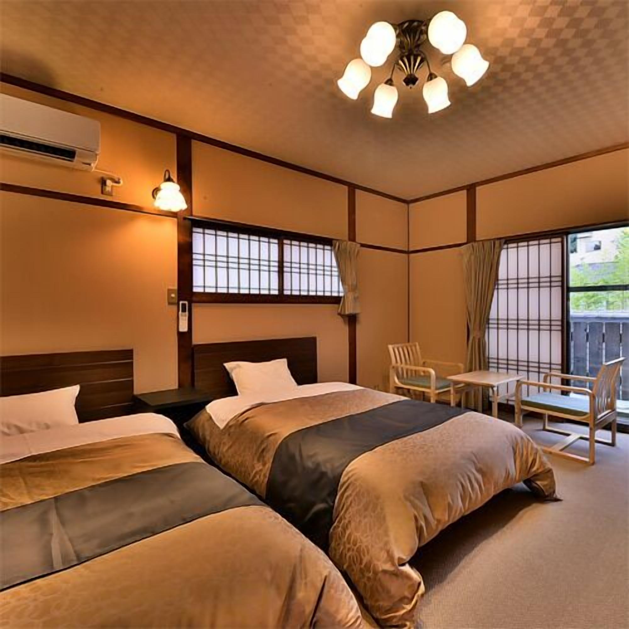 A hotel room with two beds, a small table, chairs, a window with curtains, and a ceiling light fixture.