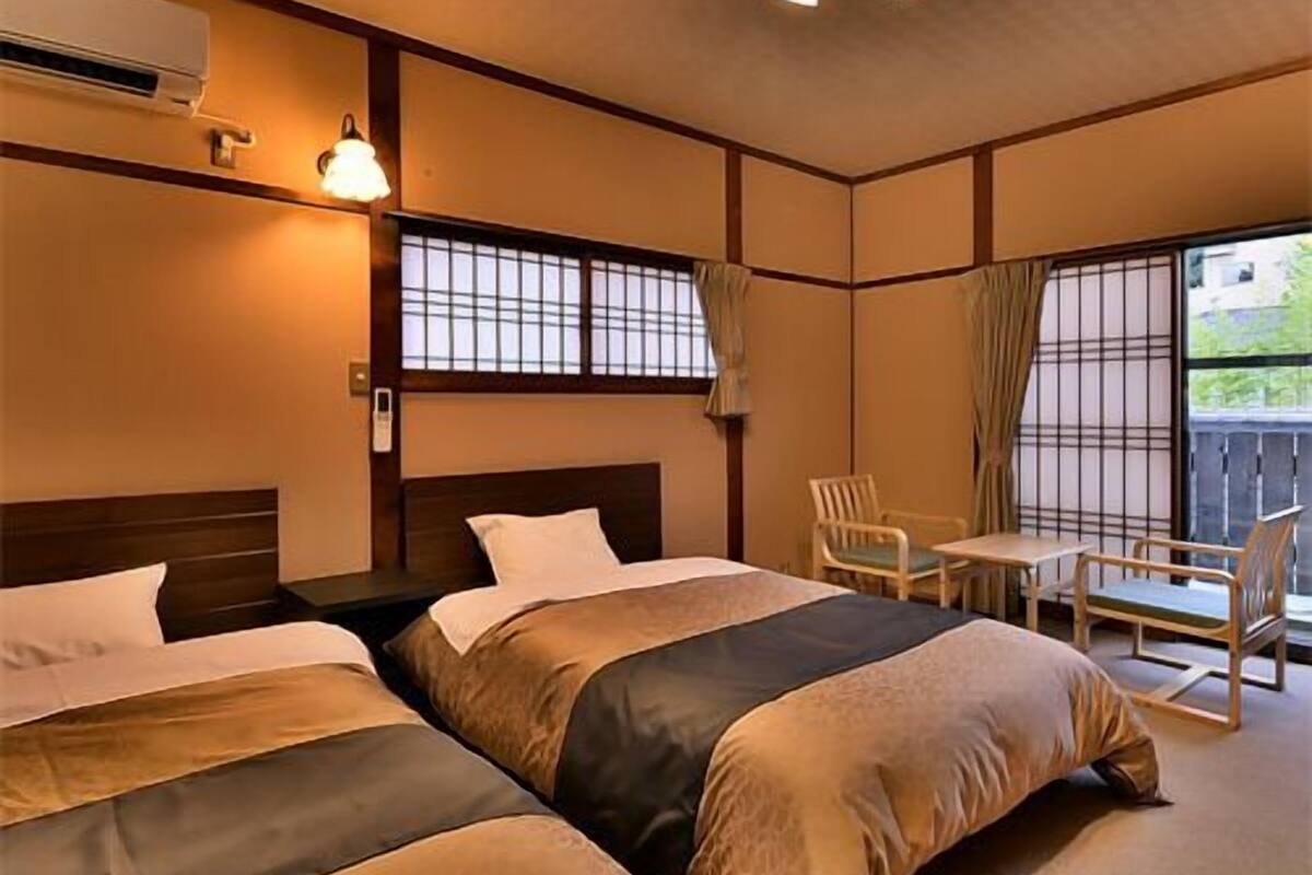 A hotel room with two beds, a small table, chairs, a window with curtains, and a ceiling light fixture.