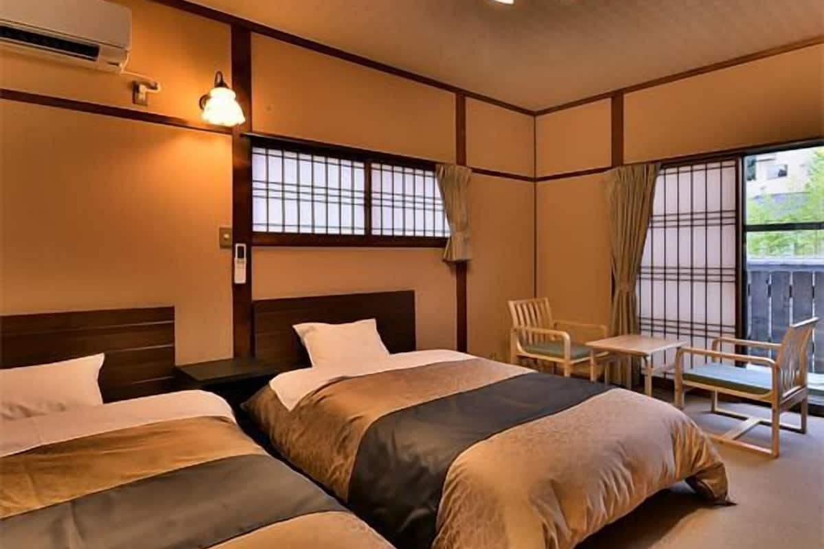 A hotel room with two beds, a small table, chairs, a window with curtains, and a ceiling light fixture.