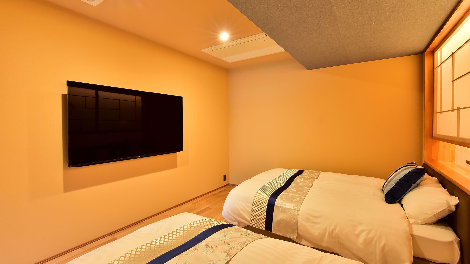 A hotel room with a flat-screen TV mounted on the wall, two beds with patterned bedding, and a wooden headboard.