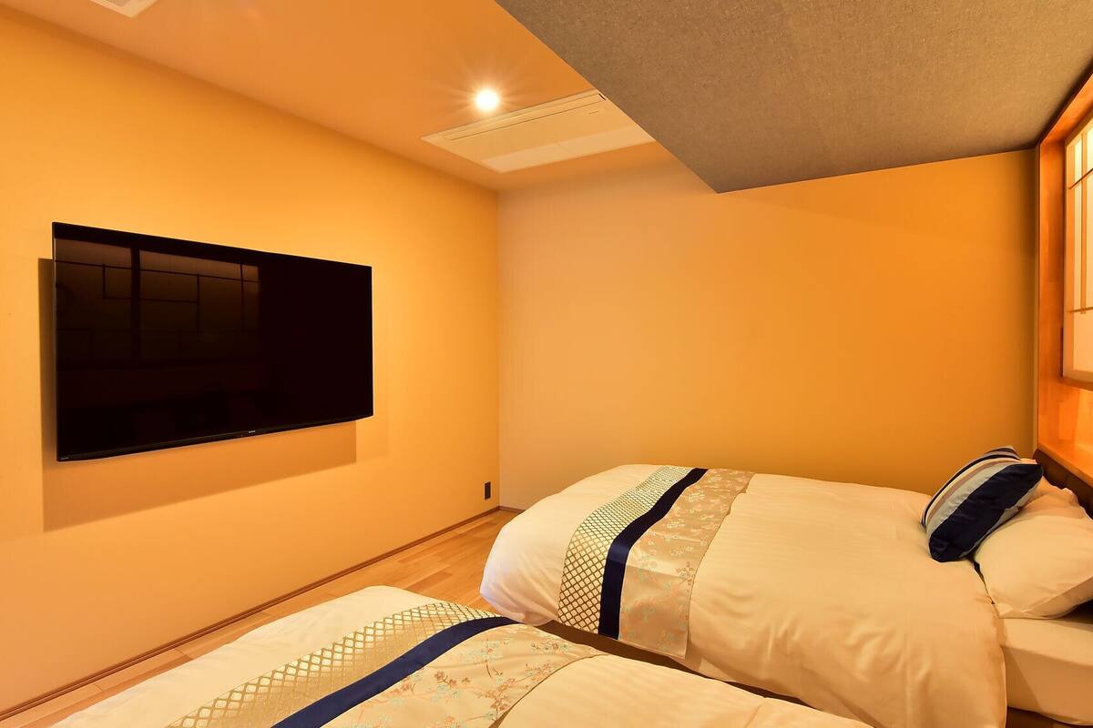 A hotel room with a flat-screen TV mounted on the wall, two beds with patterned bedding, and a wooden headboard.