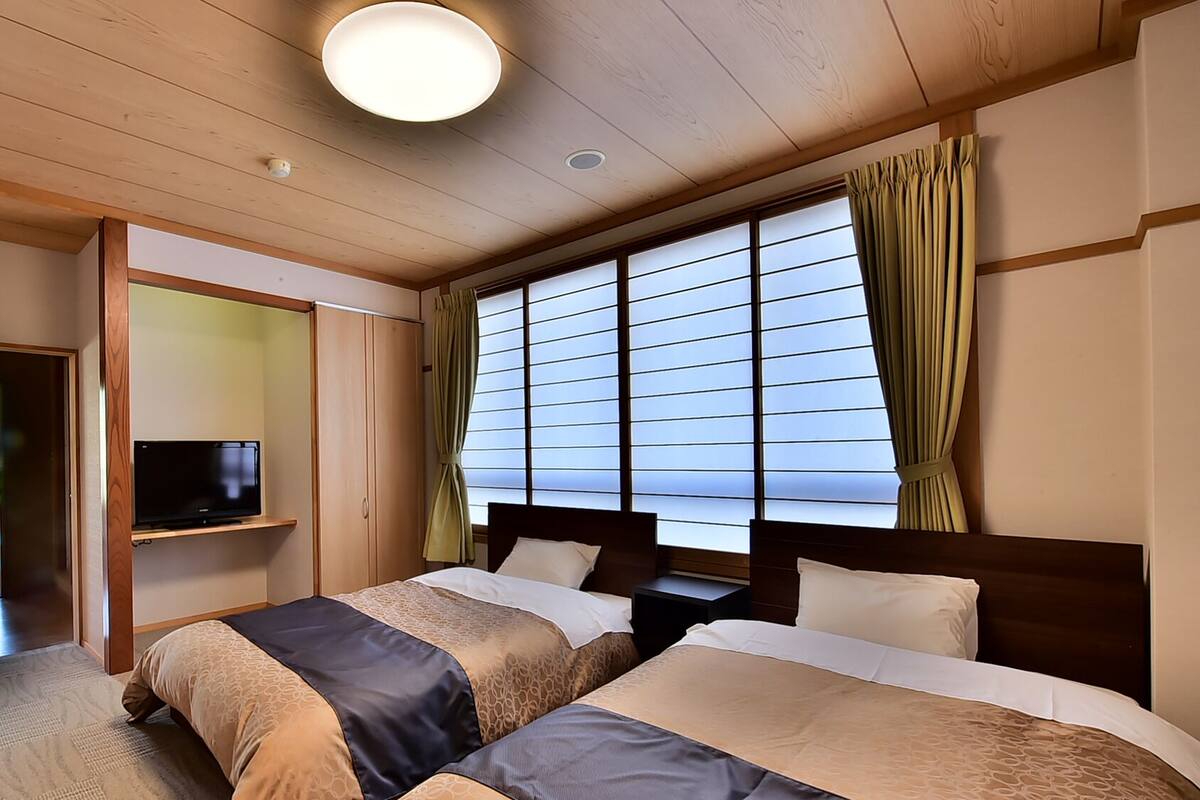 A hotel room with two beds, a TV on a shelf, a window with curtains, and a wooden ceiling.