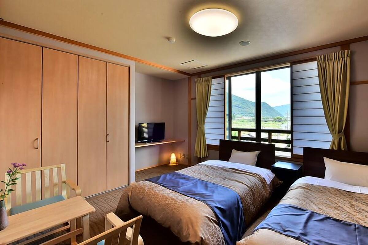 A hotel room with two beds, a desk, a TV, and a window with curtains.