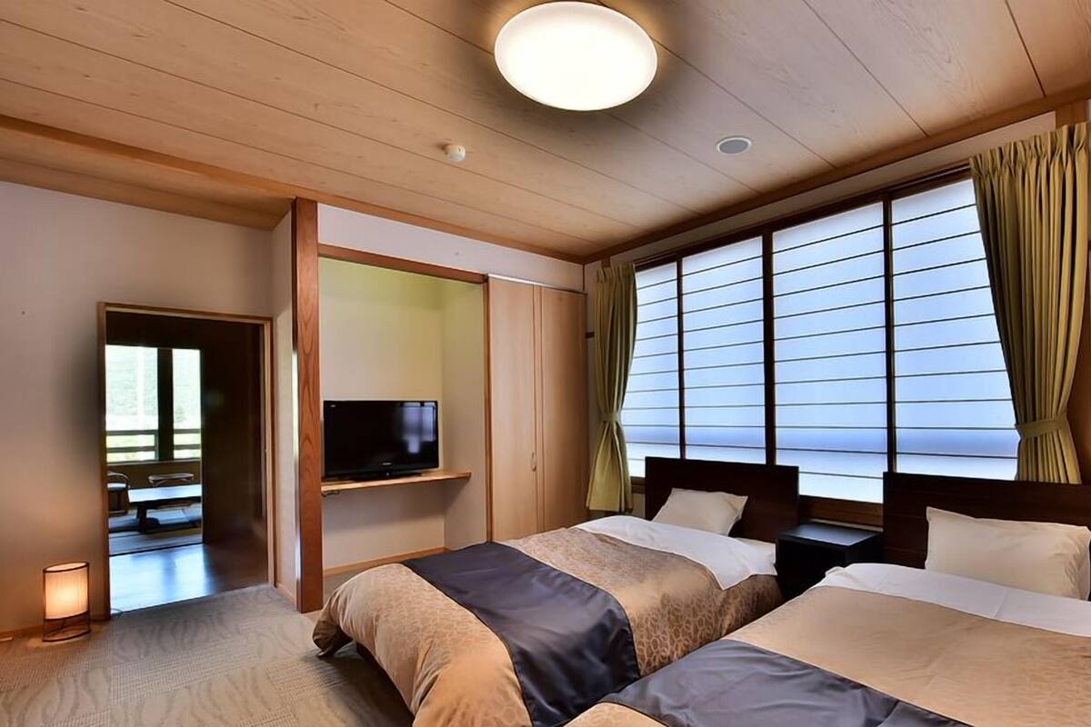 A hotel room with two beds, a television on a shelf, a window with curtains, and a ceiling light.
