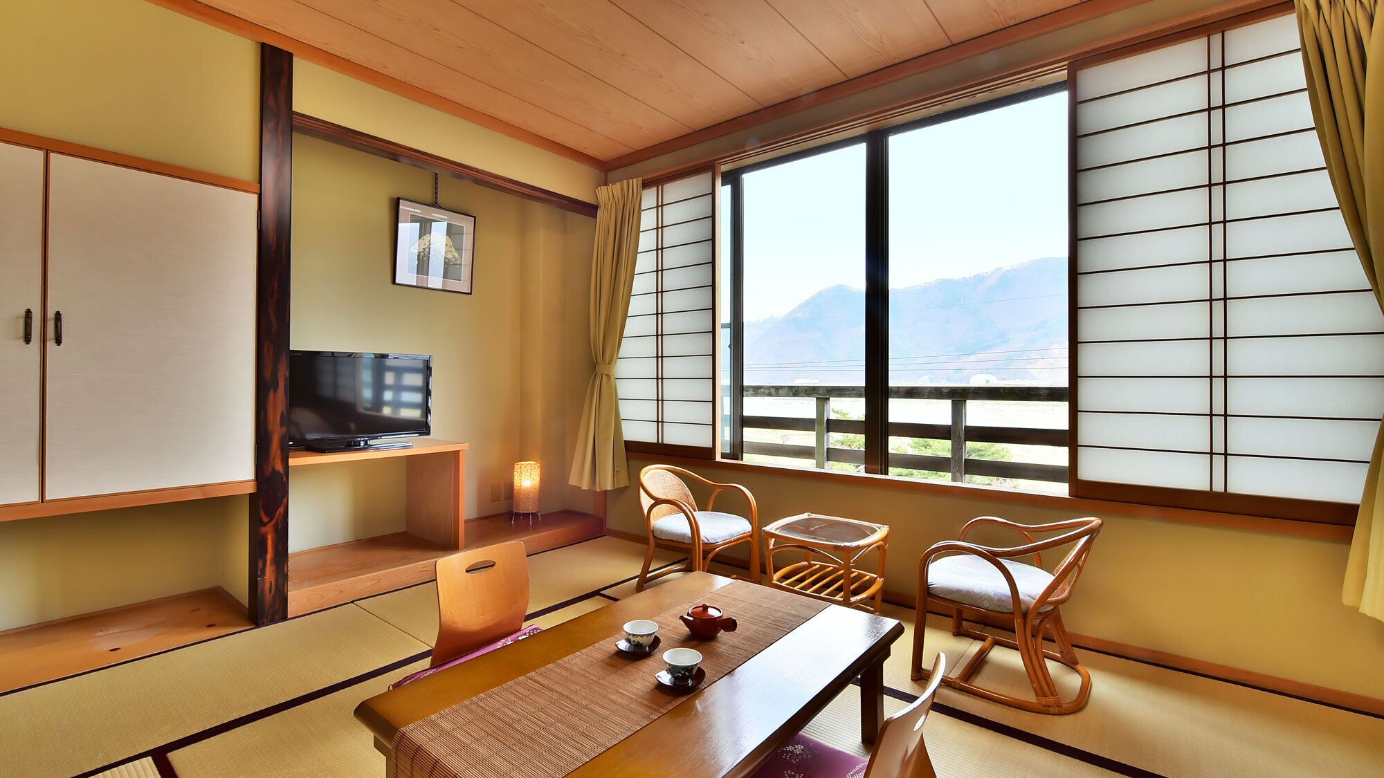 A traditional Japanese room with tatami flooring, a television, a table with tea set, and a view of mountains through shoji screens.