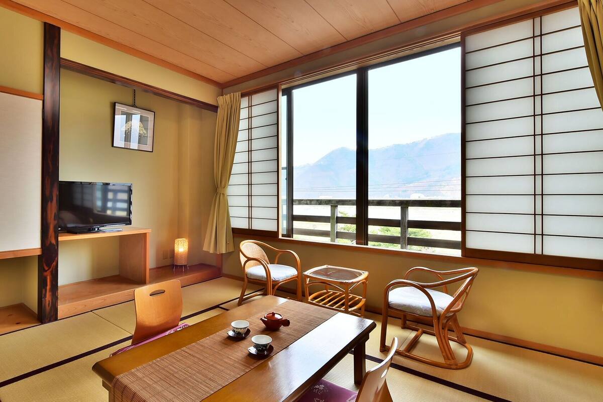 A traditional Japanese room with tatami flooring, a television, a table with tea set, and a view of mountains through shoji screens.