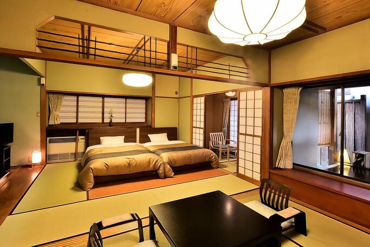 A traditional Japanese-style room with tatami flooring, a low bed, a table with chairs, and a view through a shoji screen.