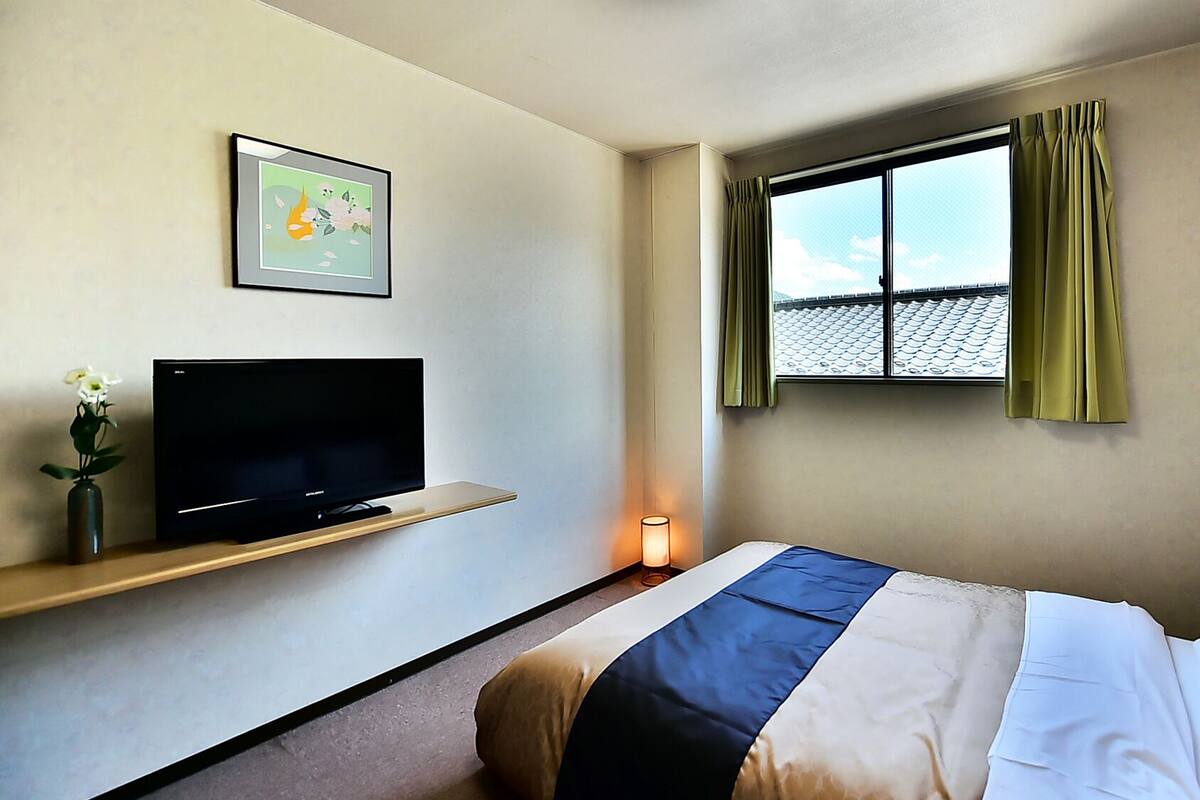A hotel room with a bed, a TV on a shelf, a window with curtains, and a wall-mounted picture.