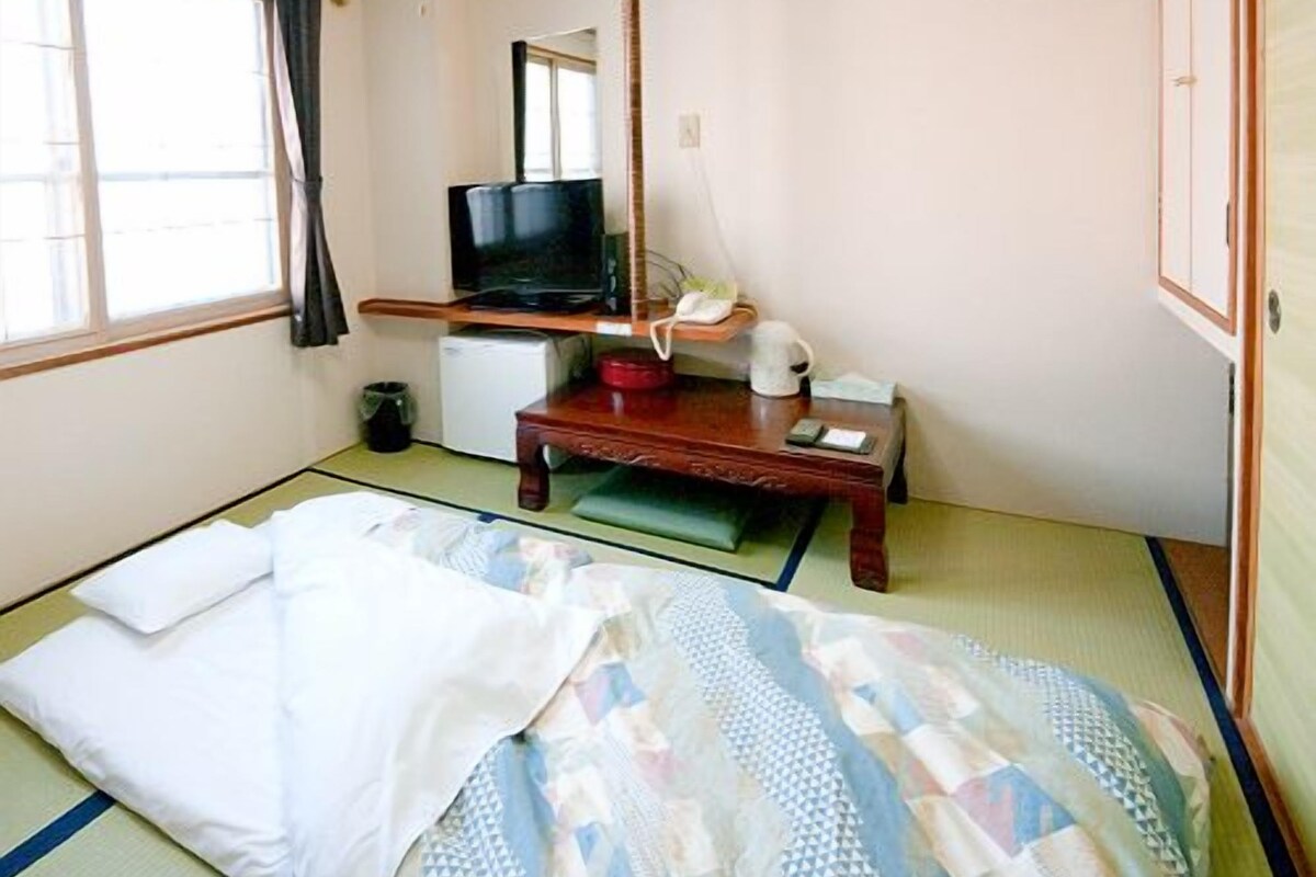 Una habitación tradicional japonesa con suelo de tatami, un escritorio de madera bajo, un televisor y una cama futón.